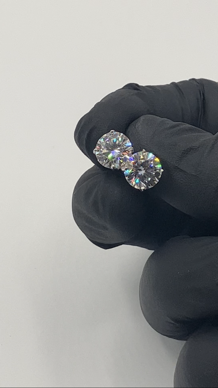 A pair of silver moissanite stud earrings held by a gloved hand, displaying their shine and color.