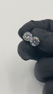 A pair of silver moissanite stud earrings held by a gloved hand, displaying their shine and color.